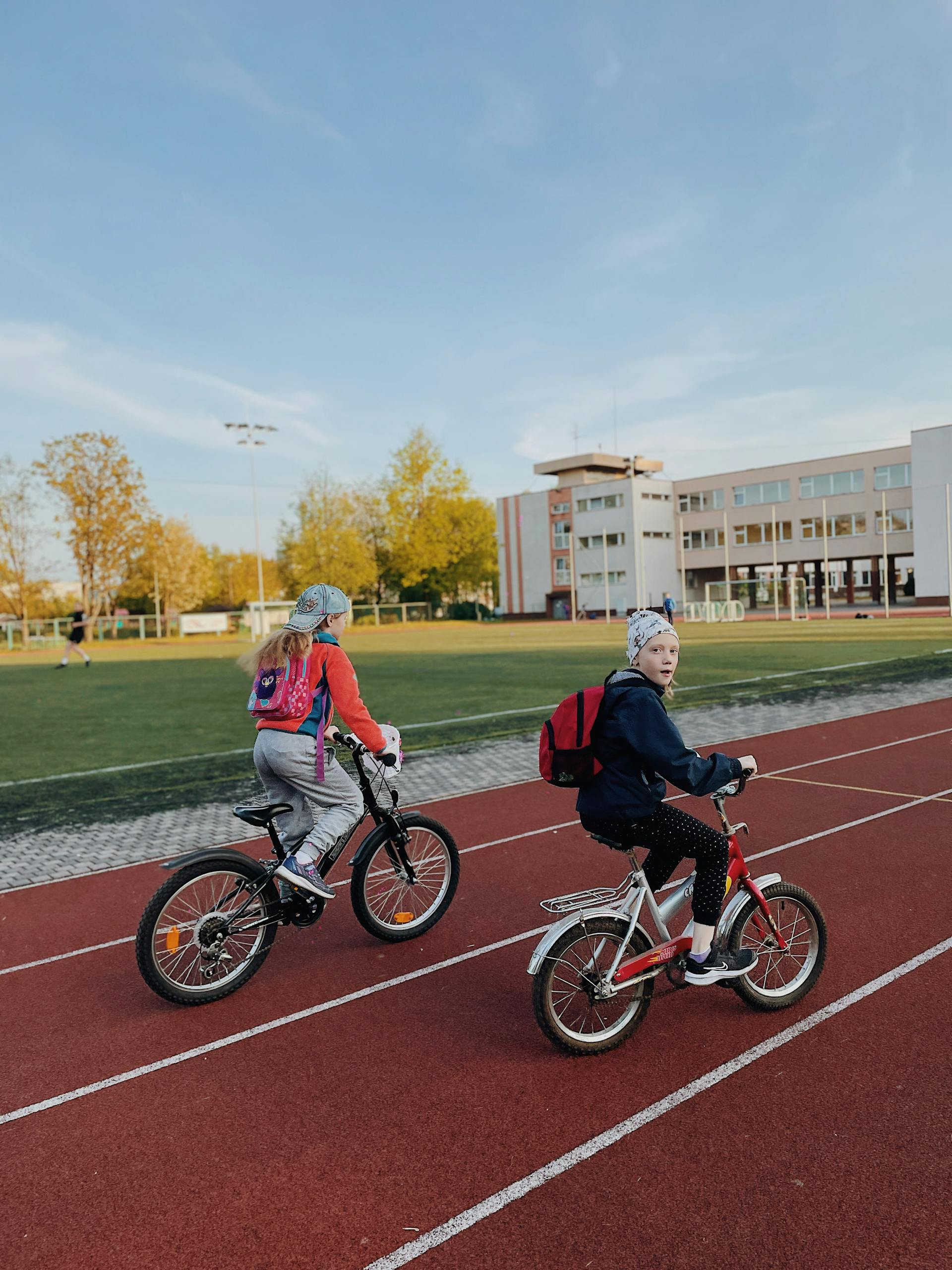 Kids riding bicycles on a school track in Rīga, Latvia, with autumn trees and a school building in the background.